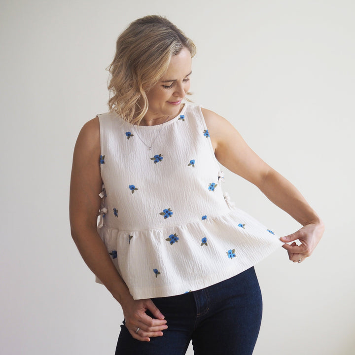 A woman wearing a sleeveless white textured top with small blue floral prints, paired with dark blue jeans, standing against a plain light background.