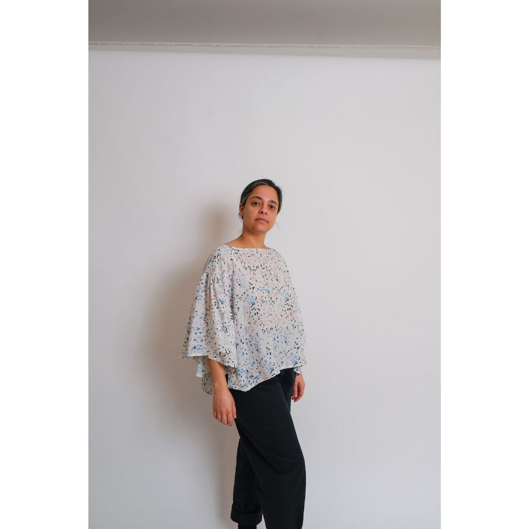 Person wearing a patterned blouse and black pants against a plain white wall