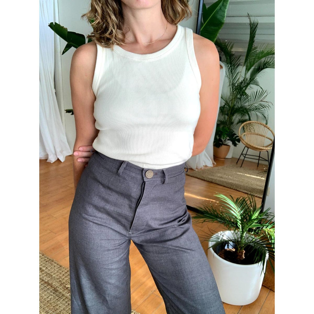 Person wearing a white sleeveless top and gray pants in a room with plants and furniture.