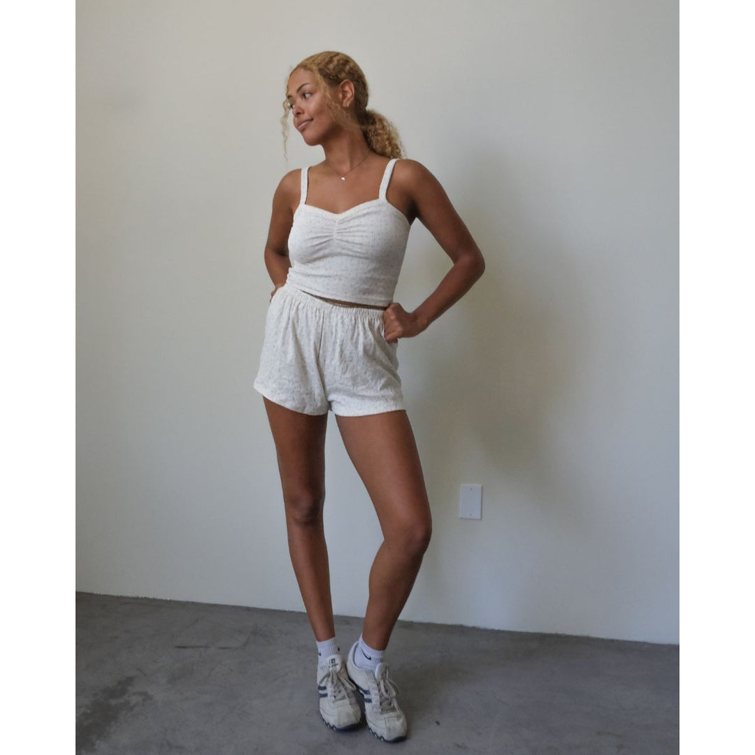Woman wearing a white tank top and shorts standing against a plain wall.