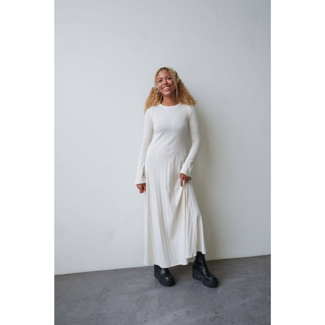 Woman wearing a long white dress standing against a plain wall.