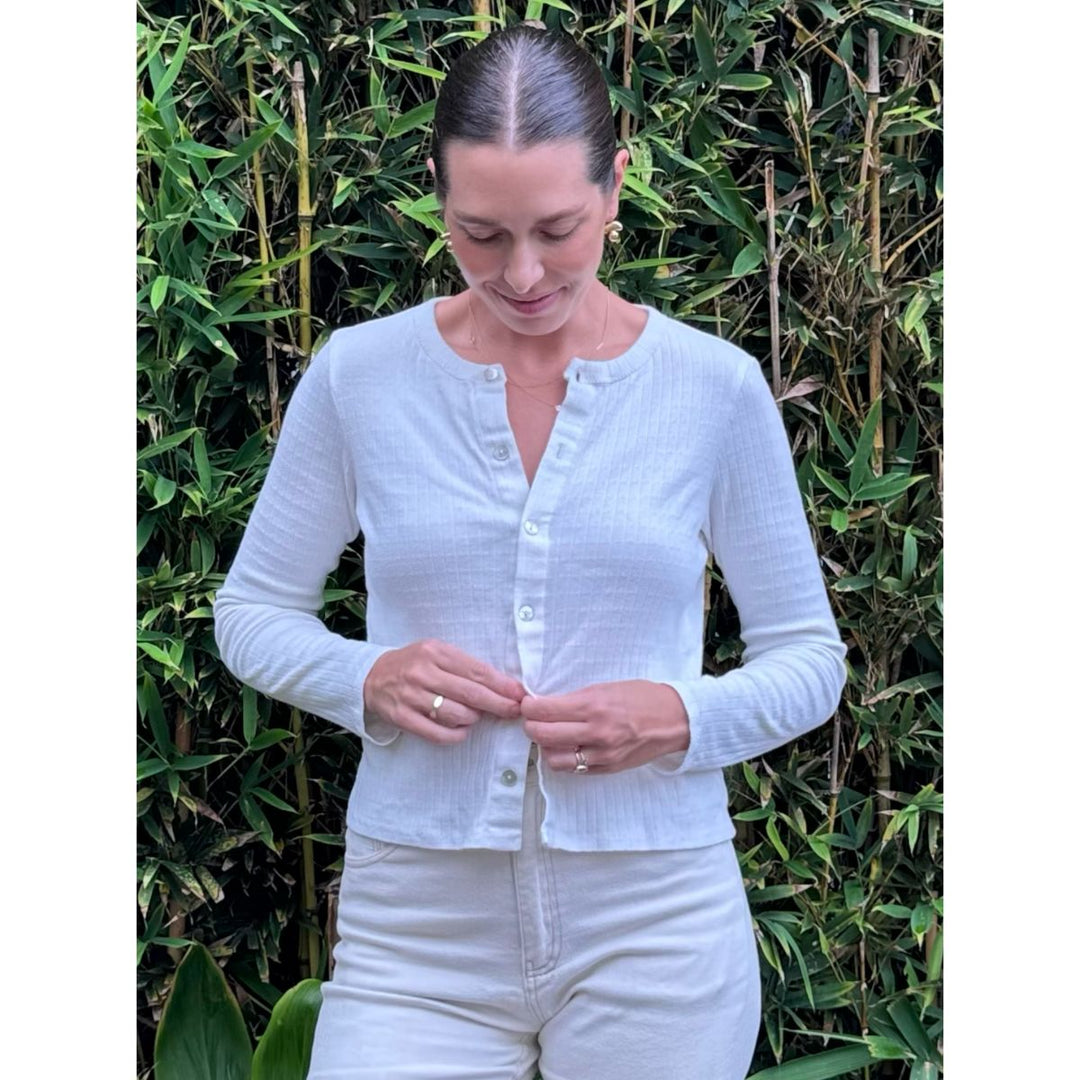 Woman wearing a light blue cardigan and white pants standing in front of green foliage.