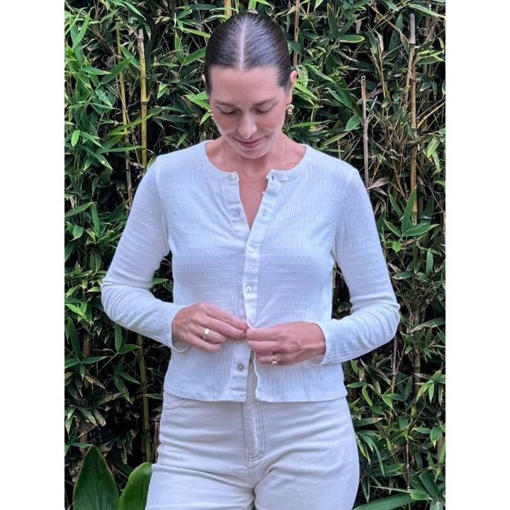 Woman wearing a light blue cardigan and white pants standing in front of green foliage.