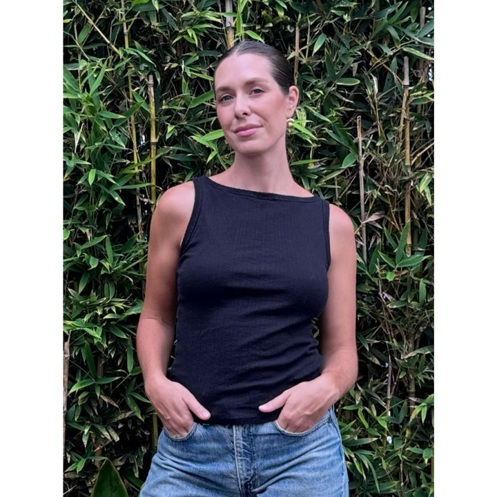 Woman wearing a black tank top and blue jeans standing against a green leafy background