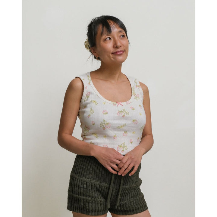 Woman wearing a white sleeveless top with floral patterns and green shorts against a plain background