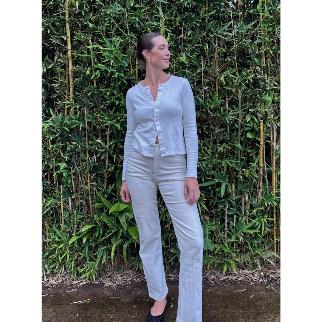 Woman wearing a light blue cardigan and white pants standing in front of a bamboo wall.