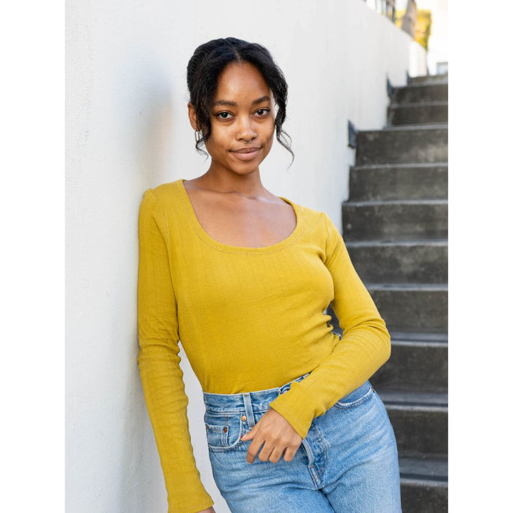 Woman wearing a yellow long-sleeve top and blue jeans standing against a white wall with steps.