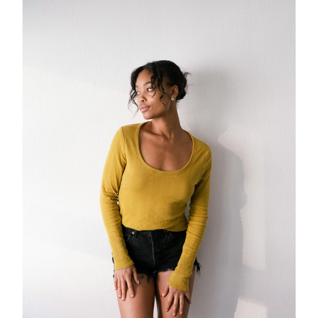 Woman wearing a mustard yellow long-sleeve top against a white wall