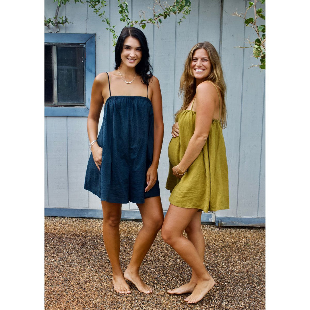 Two women standing outdoors wearing sleeveless dresses, one blue and one green, against a wooden wall.