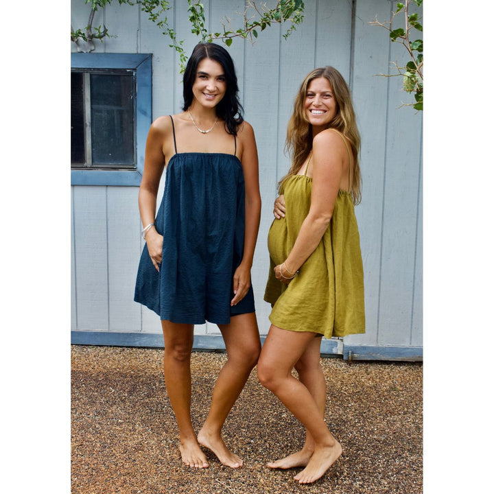Two women standing outdoors wearing sleeveless dresses, one blue and one green, against a wooden wall.