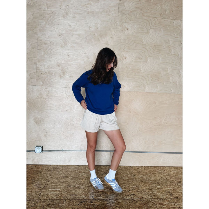 Person wearing a blue sweatshirt and white shorts standing against a beige wall.