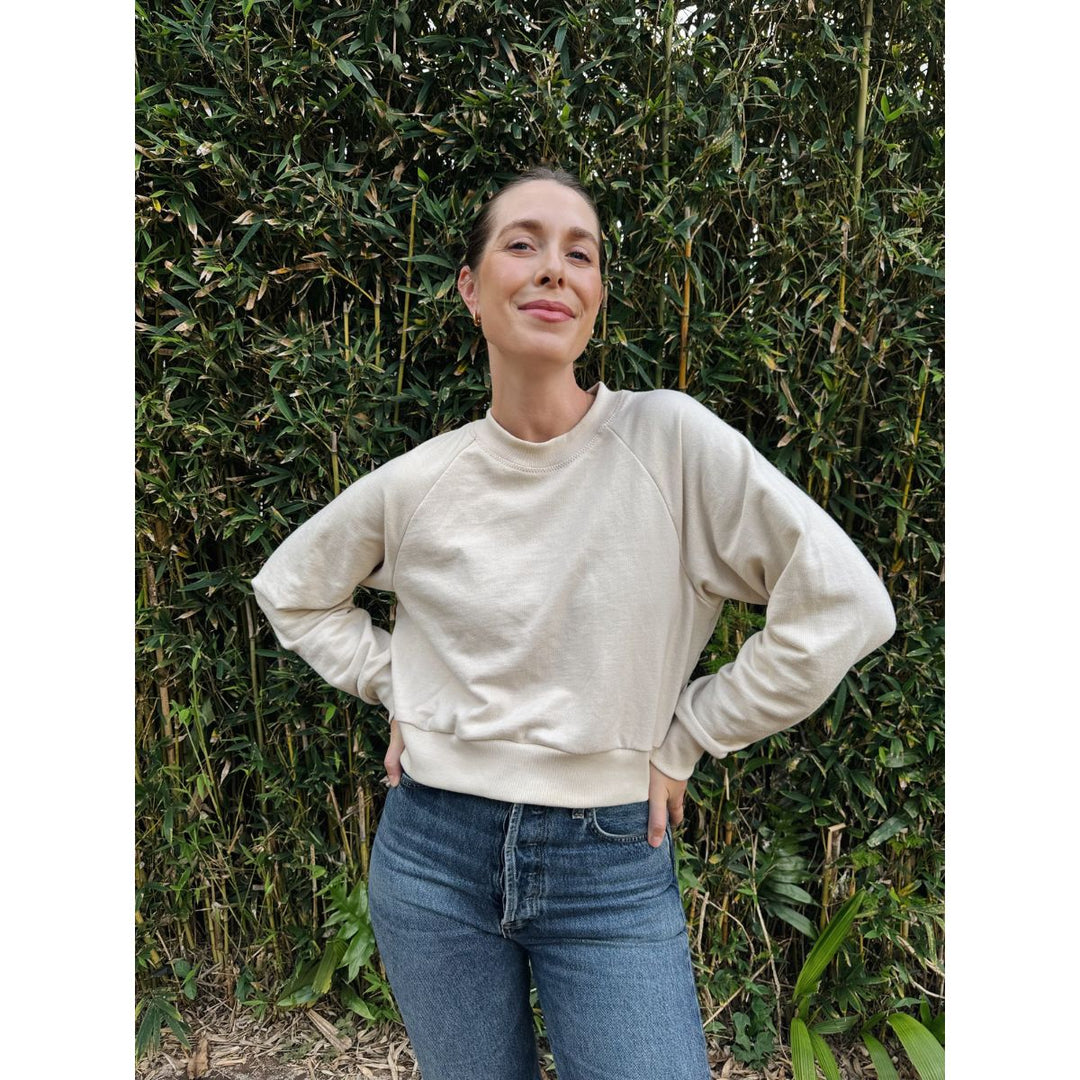 Woman wearing a beige sweater and blue jeans standing against a green bush background