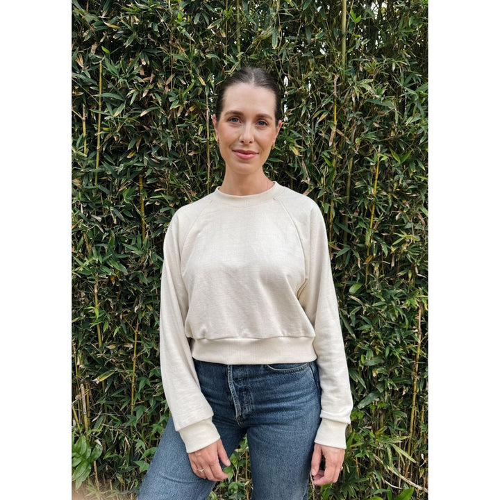 Woman wearing a beige sweater and blue jeans standing in front of a bamboo wall.