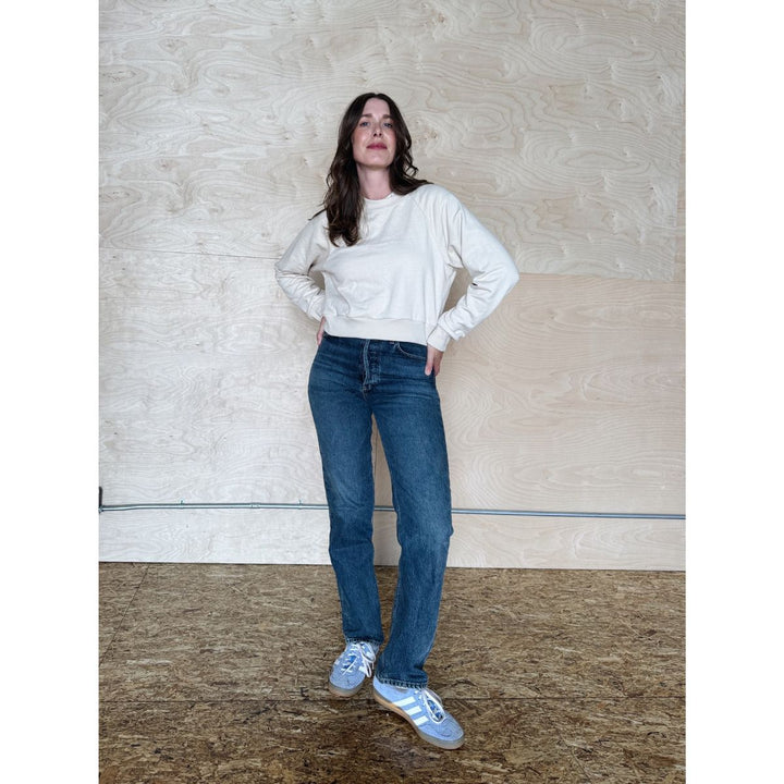 Woman wearing a white sweater and blue jeans standing against a plain wall.