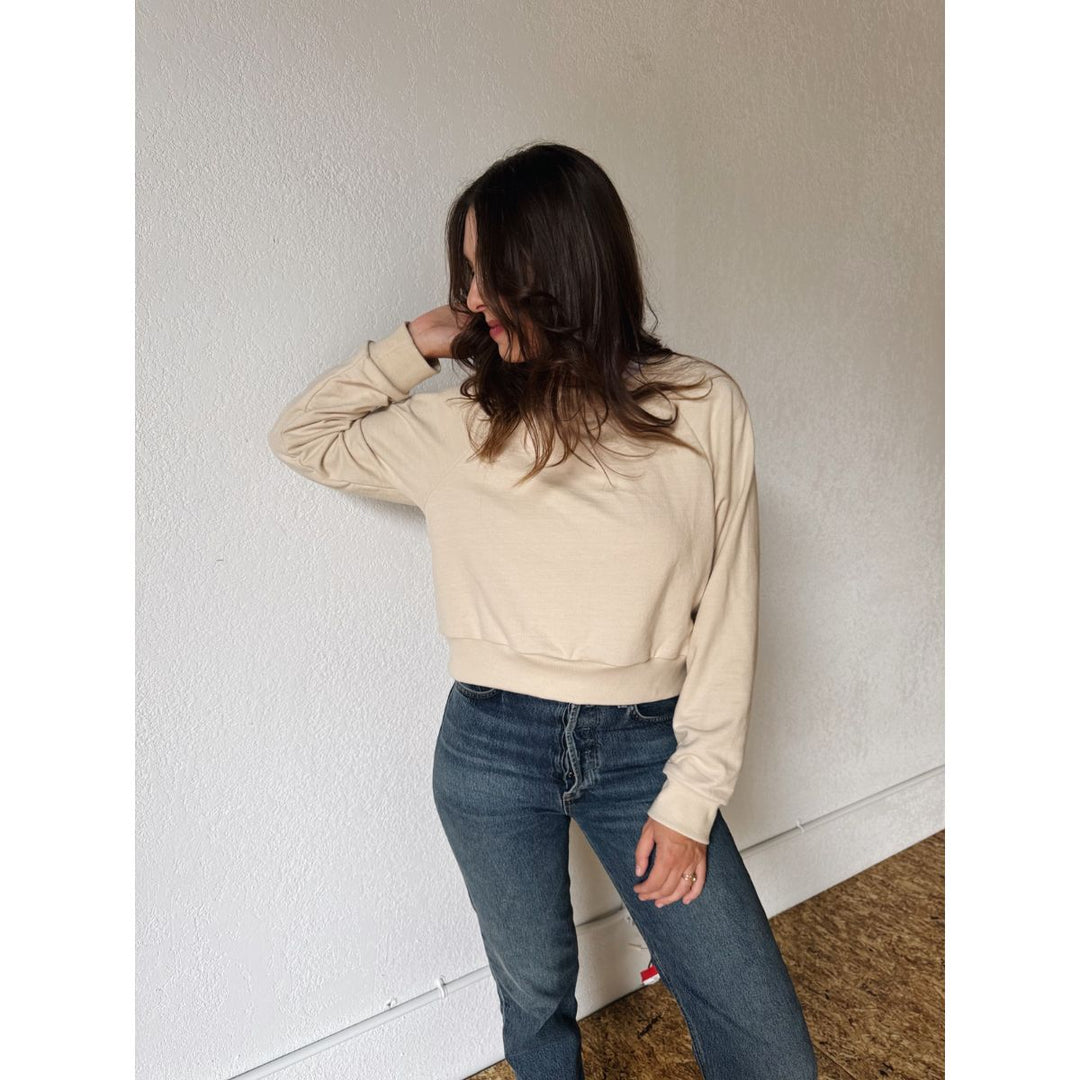Person wearing a beige sweater and blue jeans against a white wall.