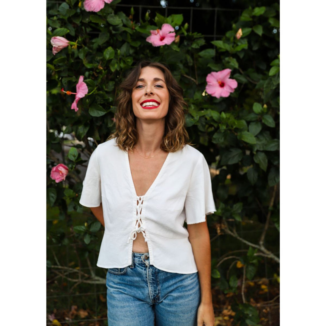 Woman wearing a white lace-up top and blue jeans standing in front of pink flowers and greenery.