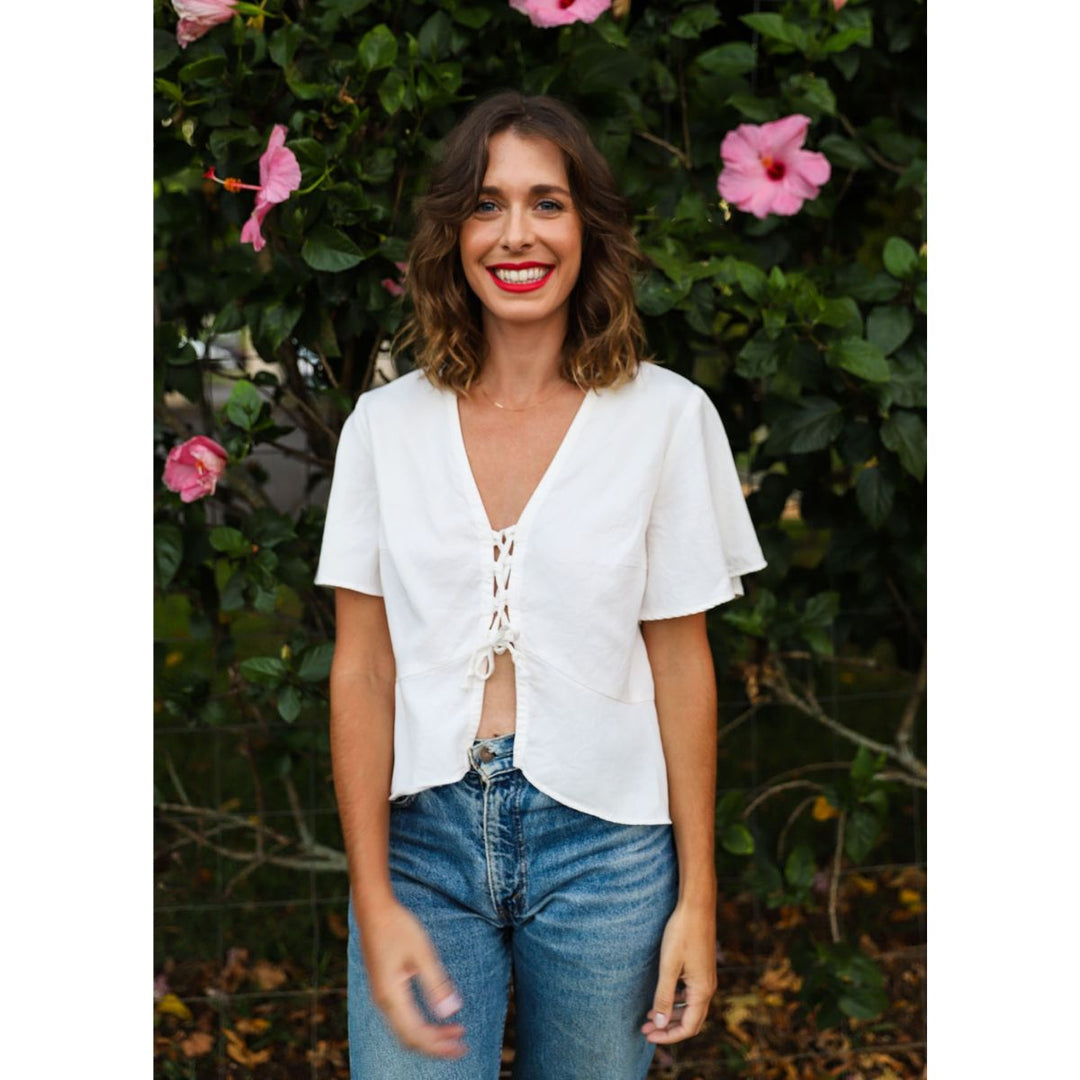Woman wearing a white blouse and blue jeans standing in front of green foliage with pink flowers.