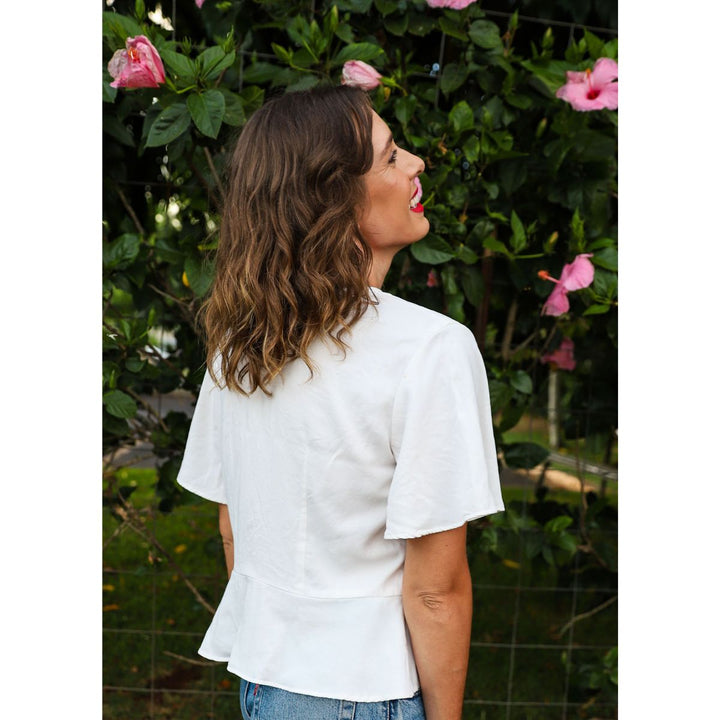 Woman wearing a white blouse standing in front of green foliage with pink flowers.