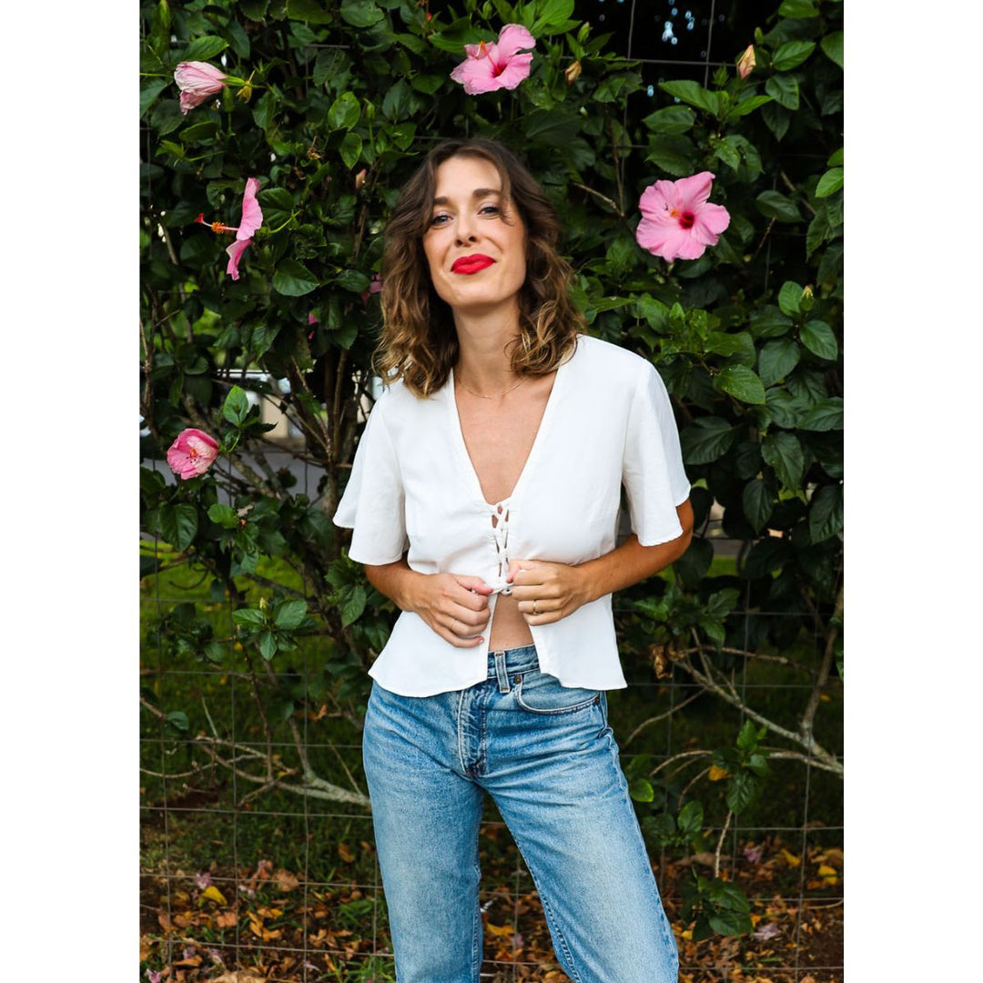 Woman in a white blouse and blue jeans standing in front of green foliage with pink flowers.