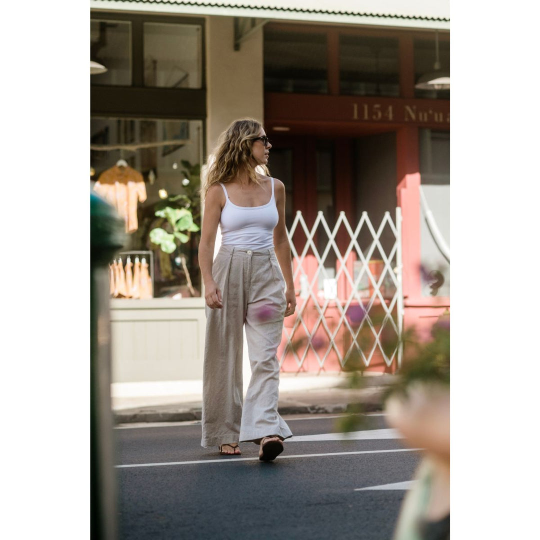 Woman walking on a street wearing a white tank top and beige pants.