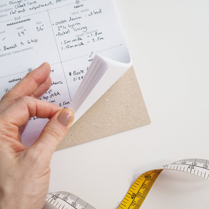Hand holding a piece of paper with measurements, next to a tape measure on a white surface.