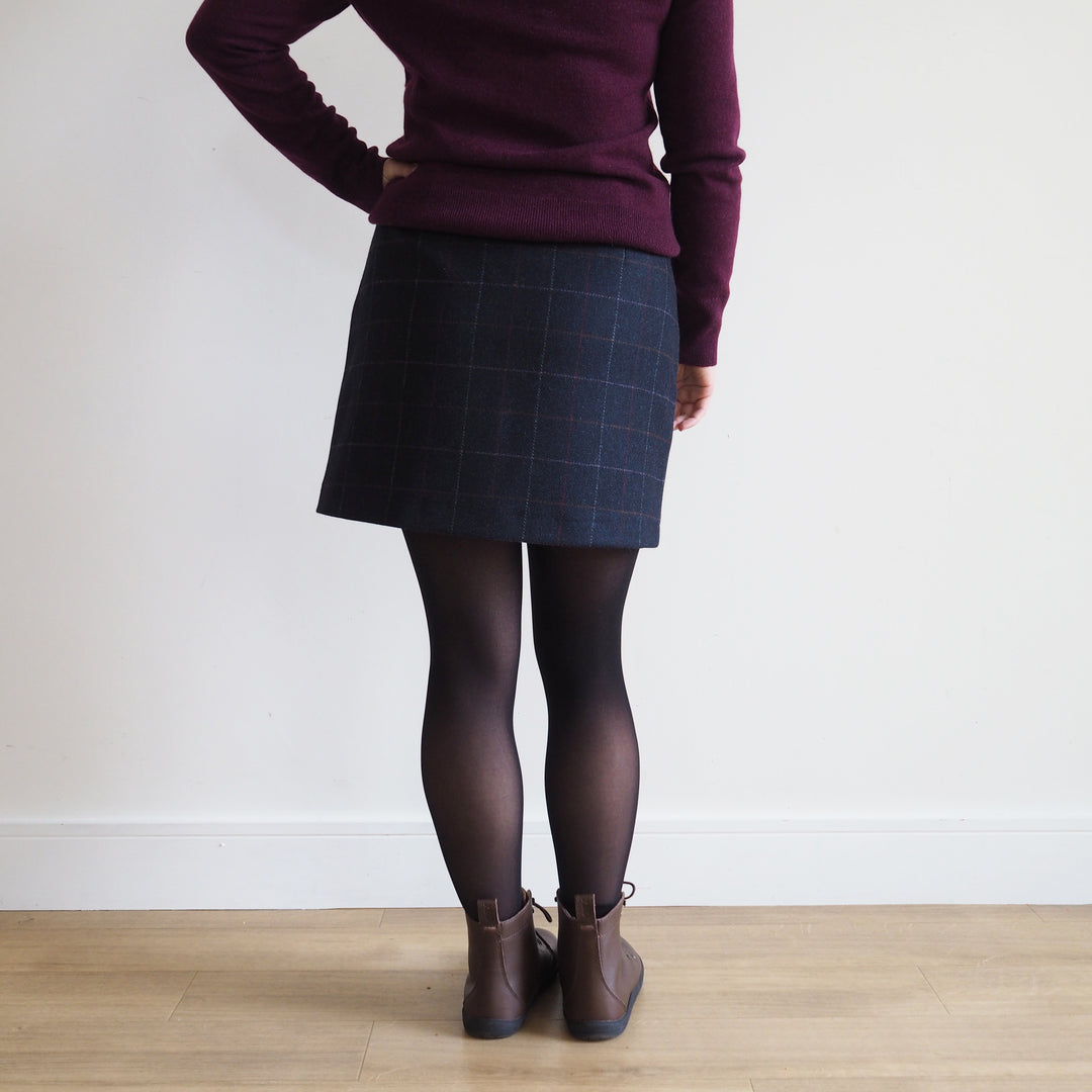 Person wearing a purple sweater, dark plaid skirt, black tights, and brown shoes on a plain background