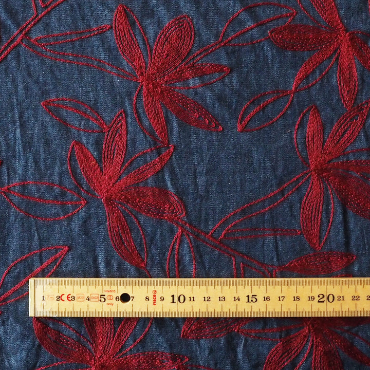 A close-up of dark blue fabric with red embroidered floral patterns, with a wooden ruler at the bottom showing scale.