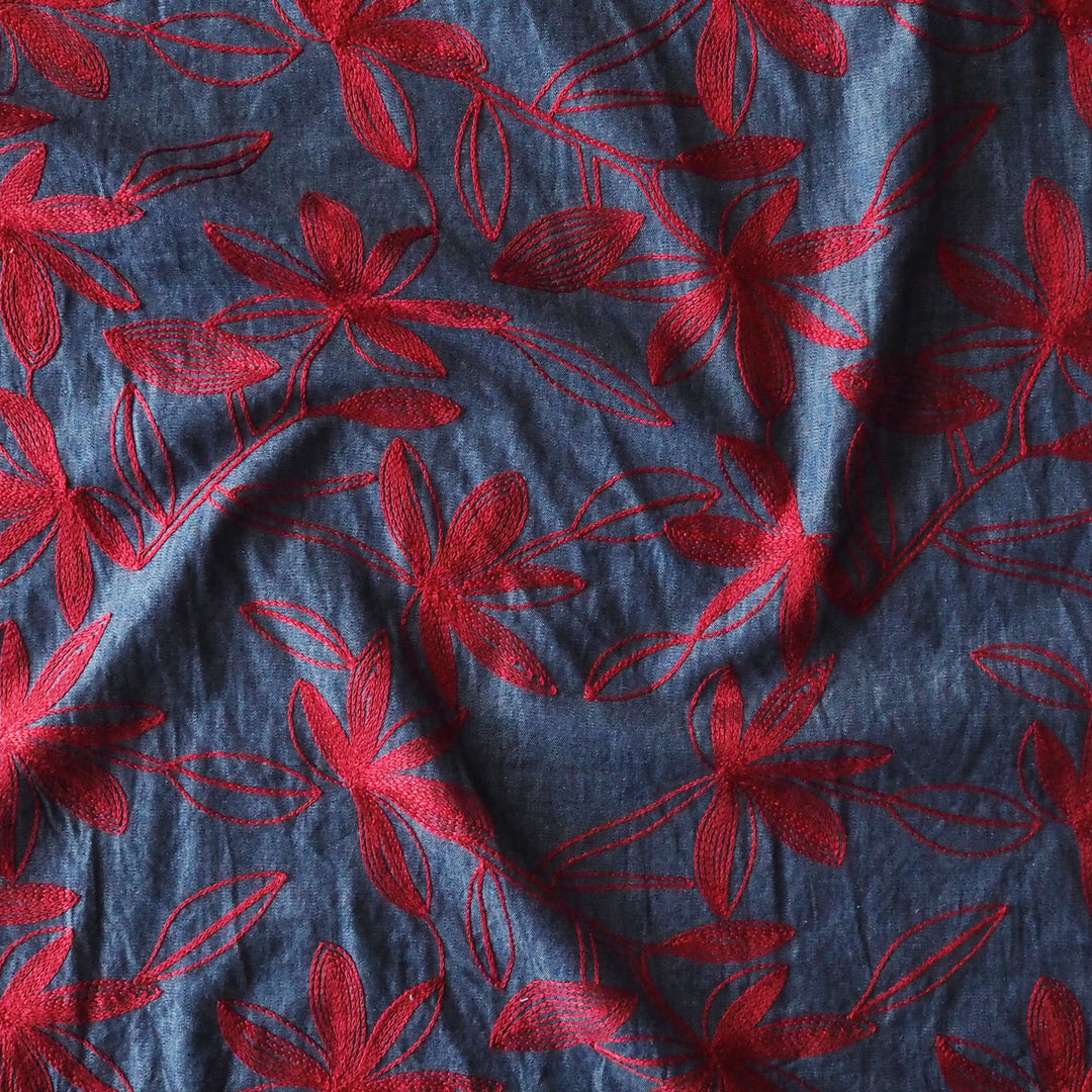 A close-up of a blue-gray fabric with a red embroidered floral pattern, shown with soft folds.