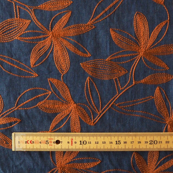 A close-up of dark blue fabric with large rust-orange floral embroidery, with a wooden ruler along the bottom edge showing measurements.