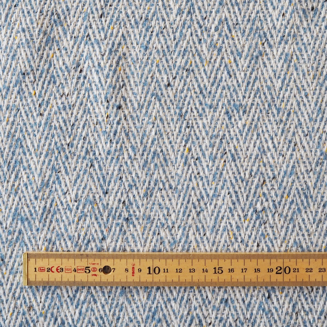A close-up of a woven fabric with a blue and white herringbone pattern, featuring small flecks of yellow and black. A wooden ruler is placed along the bottom edge to show scale.