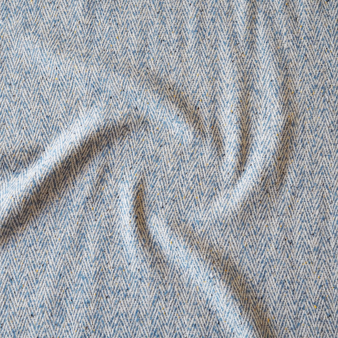 A close-up of a textured woven fabric with a herringbone pattern in light blue, beige, and gray, shown with soft folds.
