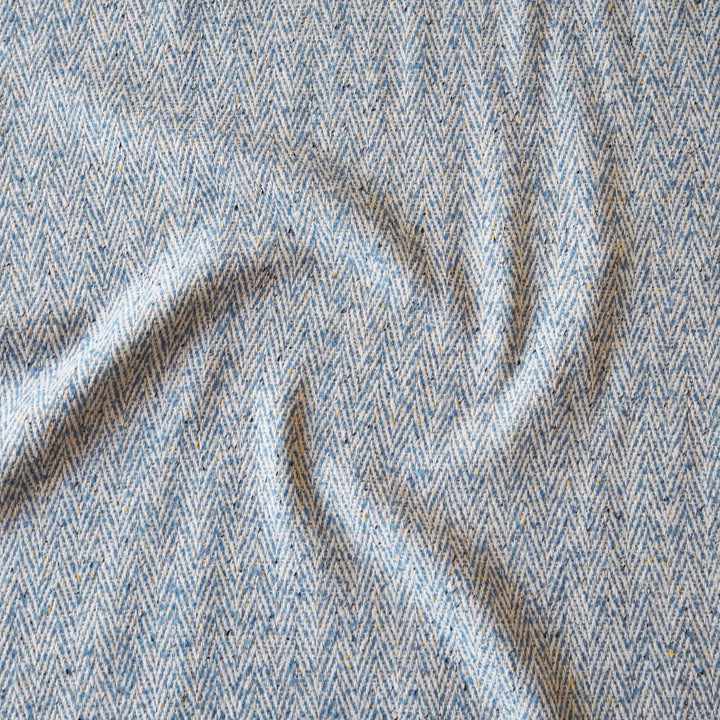 A close-up of a textured woven fabric with a herringbone pattern in light blue, beige, and gray, shown with soft folds.
