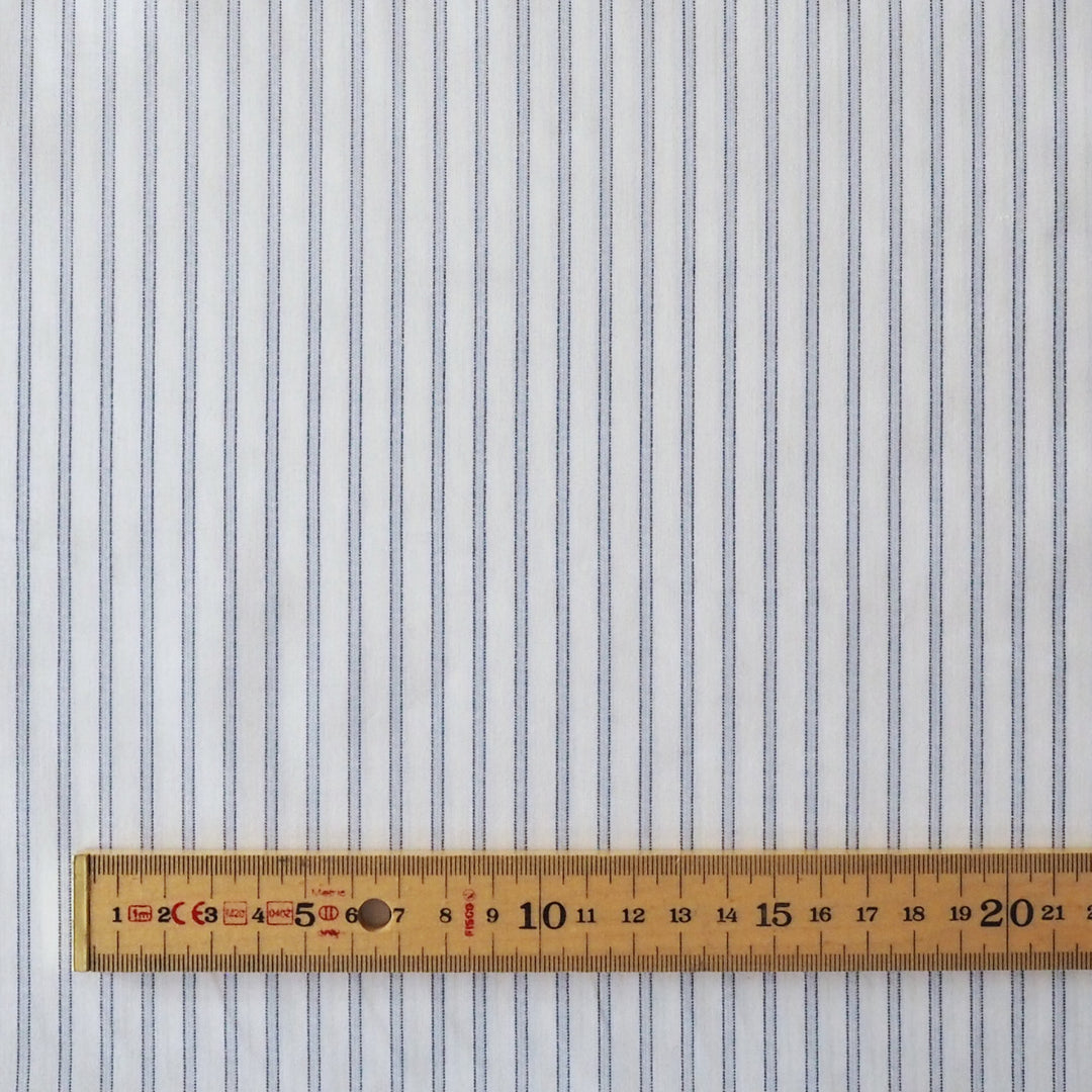 White fabric with thin vertical blue pinstripes, shown flat with a wooden ruler at the bottom for scale.