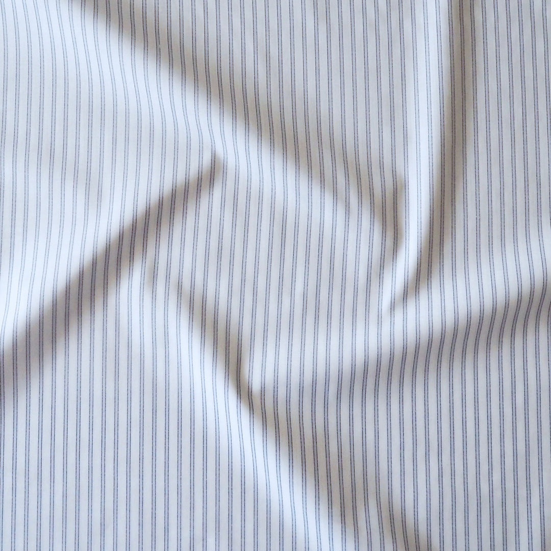A close-up of draped fabric with a fine vertical stripe pattern in navy blue on a warm off-white or cream background.