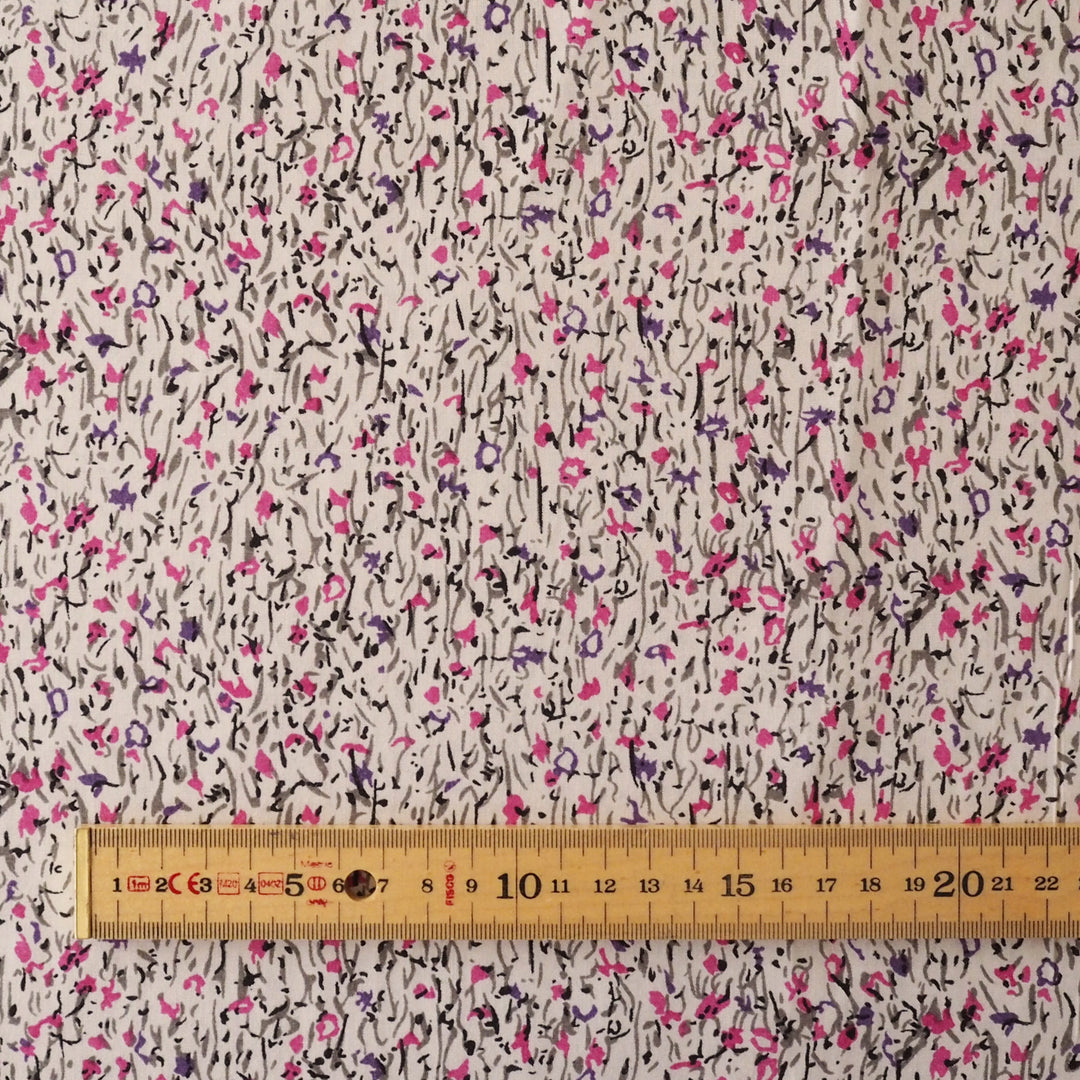 A close-up of a fabric surface with an allover abstract floral print on a light beige background, featuring black, pink, and purple scribbled flower motifs. A wooden ruler is placed along the bottom edge to show scale.