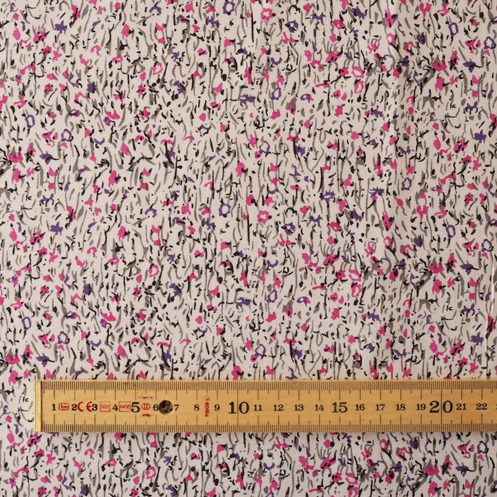 A close-up of a fabric surface with an allover abstract floral print on a light beige background, featuring black, pink, and purple scribbled flower motifs. A wooden ruler is placed along the bottom edge to show scale.