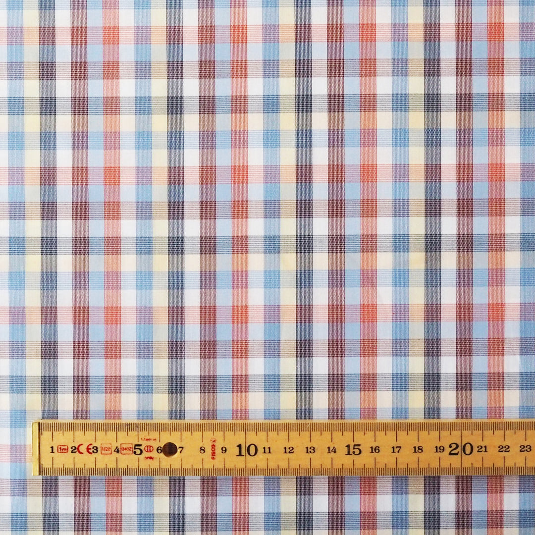 A close-up of a plaid fabric pattern with intersecting blue, red, white, and beige stripes, shown with a wooden ruler along the bottom edge for scale.
