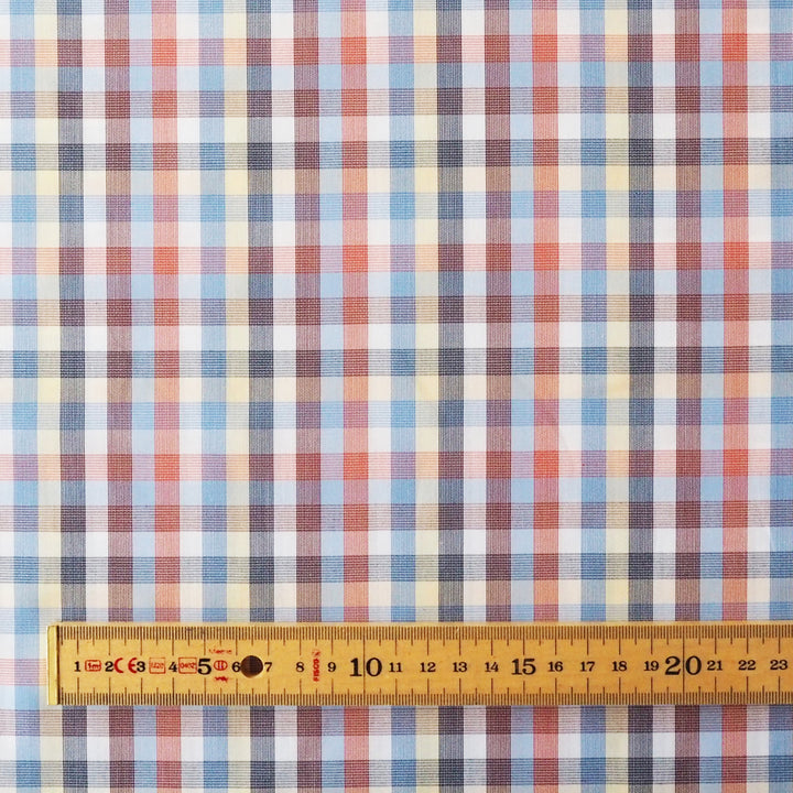 A close-up of a plaid fabric pattern with intersecting blue, red, white, and beige stripes, shown with a wooden ruler along the bottom edge for scale.