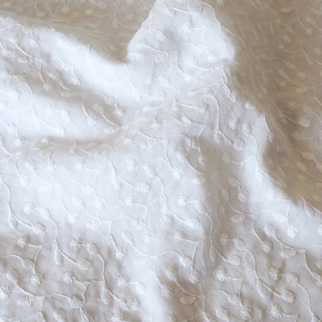A close-up of white textured fabric with a subtle floral and vine pattern, draped in soft folds.