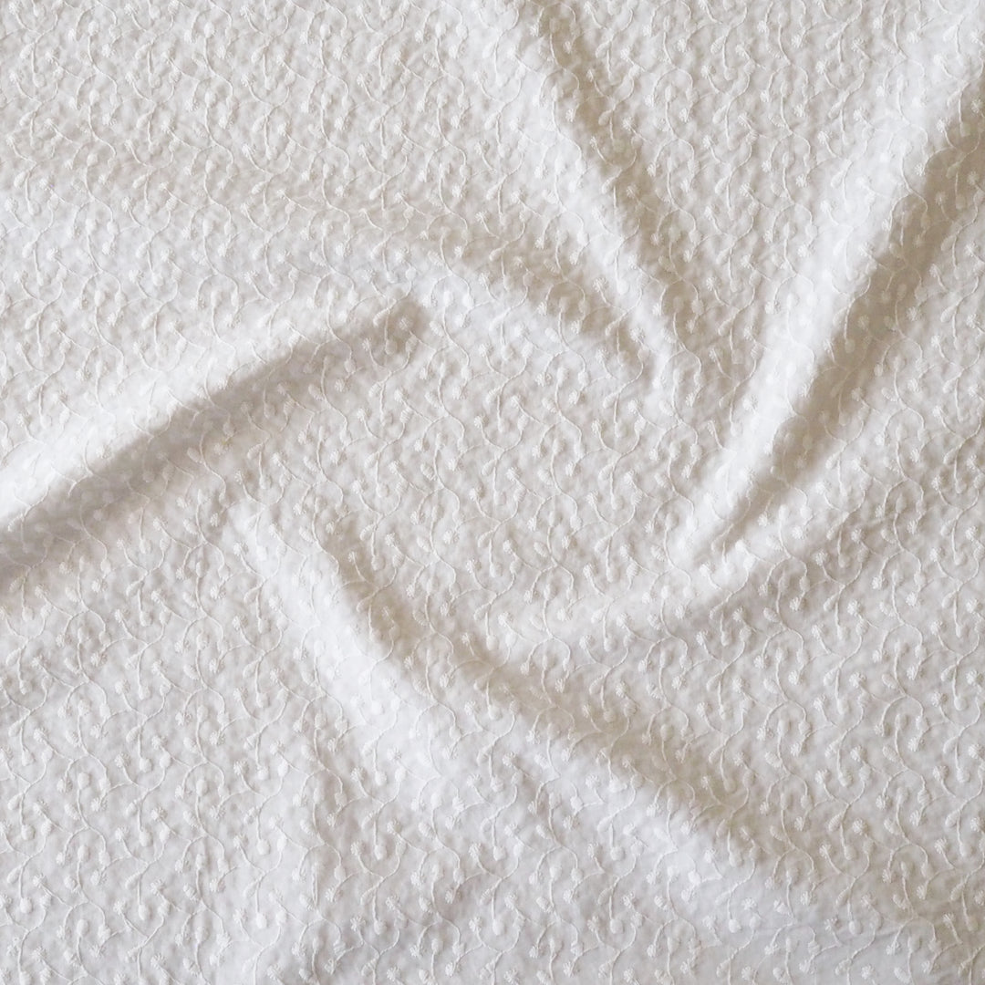 A close-up of white textured fabric with a subtle embroidered floral pattern, arranged in soft folds.