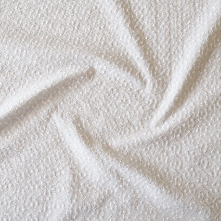 A close-up of white textured fabric with a subtle embroidered floral pattern, arranged in soft folds.