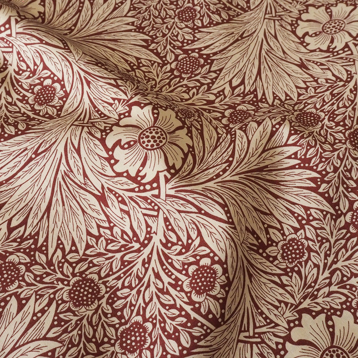 A close-up of patterned fabric with a repeating botanical design in warm beige and deep burgundy red, showing leaves, flowers, and stylised motifs.