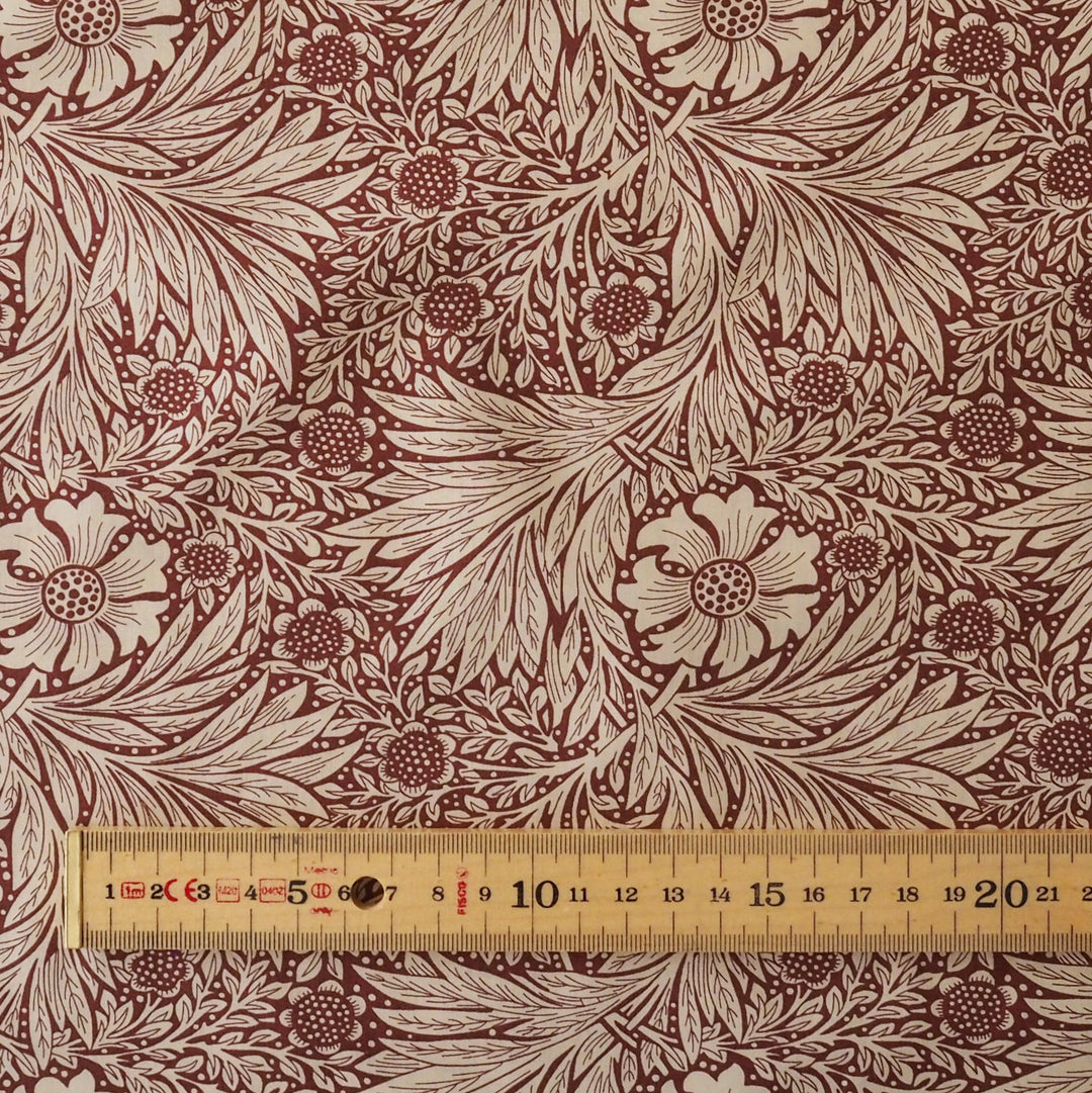 A close-up of a fabric pattern featuring a classic floral and leafy design in dark red and beige, with a wooden ruler at the bottom showing scale.