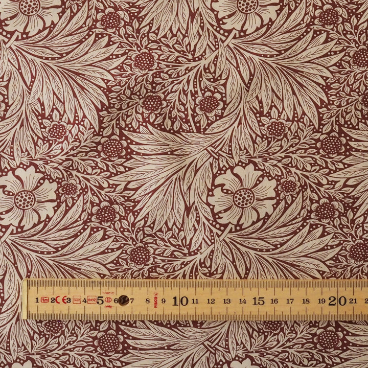 A close-up of a fabric pattern featuring a classic floral and leafy design in dark red and beige, with a wooden ruler at the bottom showing scale.