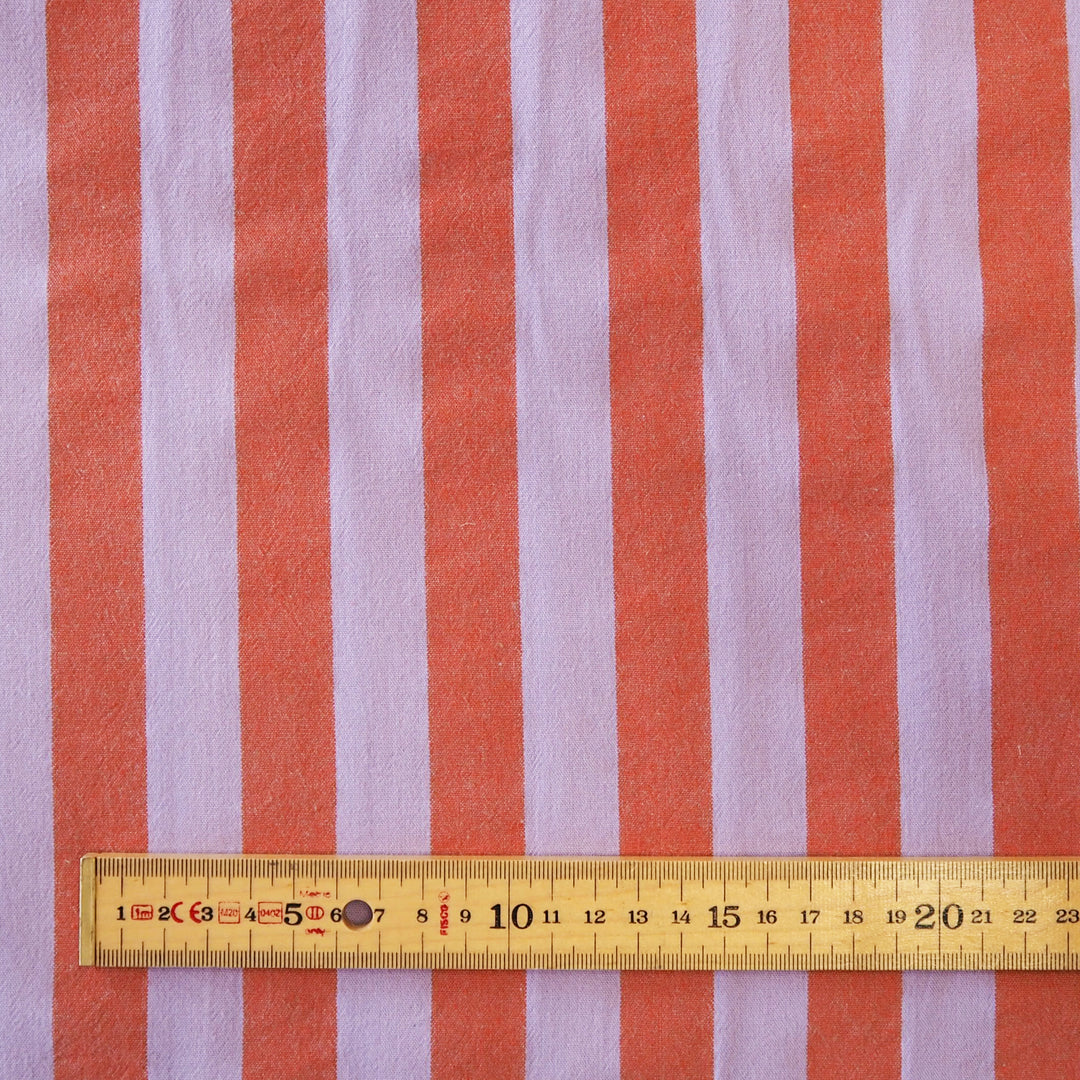 A close-up of striped fabric with alternating coral orange and light lavender vertical stripes, with a wooden ruler along the bottom edge showing the scale of the pattern.