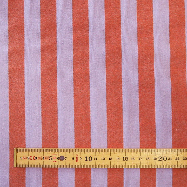 A close-up of striped fabric with alternating coral orange and light lavender vertical stripes, with a wooden ruler along the bottom edge showing the scale of the pattern.