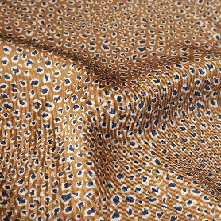 A close-up of leopard-print fabric with a warm tan base, scattered dark blue spots outlined in black, and cream borders, shown with soft folds.