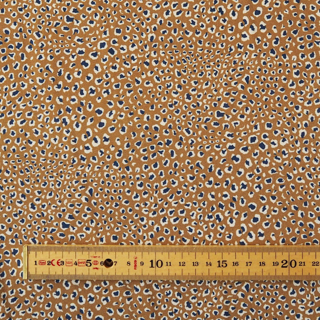 A close-up of a fabric pattern with a warm tan background covered in small irregular spots in navy blue, cream, and light brown, with a wooden ruler along the bottom edge for scale.