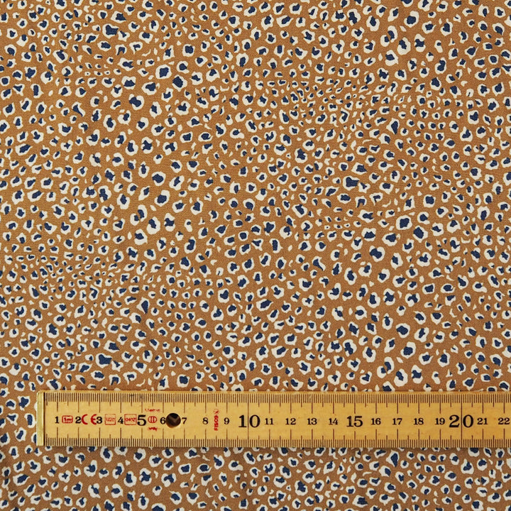A close-up of a fabric pattern with a warm tan background covered in small irregular spots in navy blue, cream, and light brown, with a wooden ruler along the bottom edge for scale.