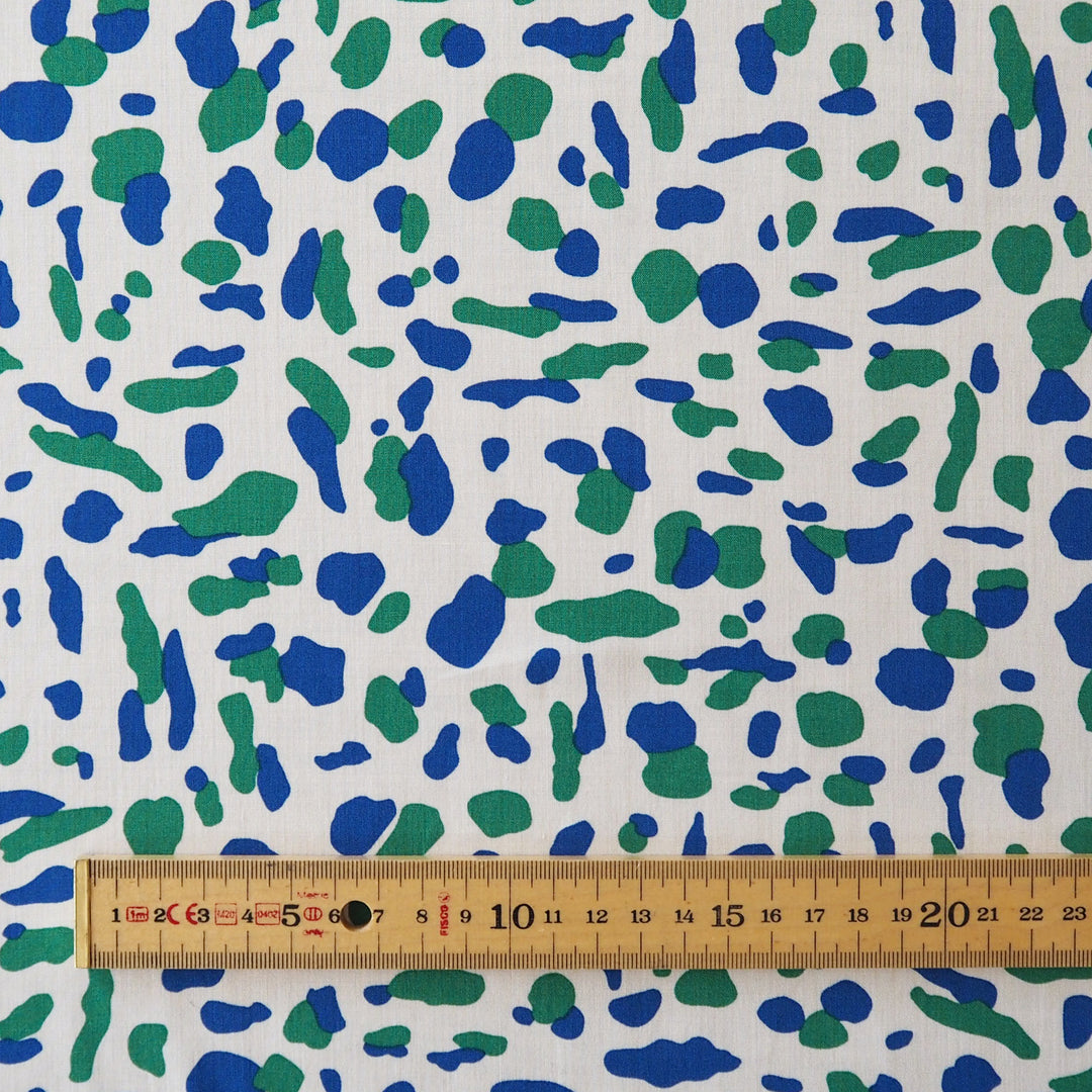 White fabric with an abstract blue and green spotted pattern, shown with a wooden ruler at the bottom for scale.